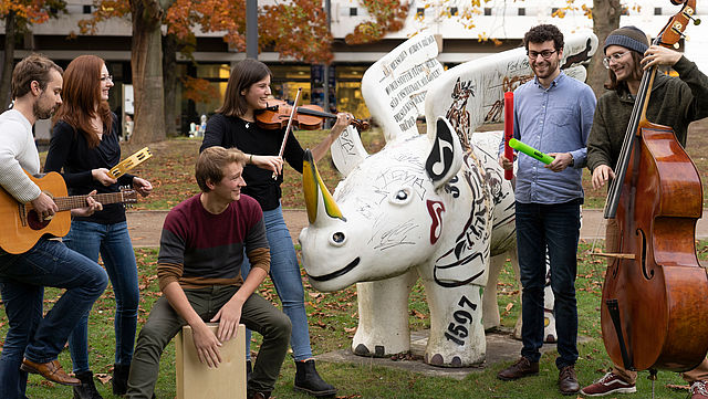 Eine Gruppe Studierender musiziert im Park der Universität. In ihrer Mitte steht ein geflügeltes Nashorn (Statue), das Symbol des Konzerthauses Dortmund.