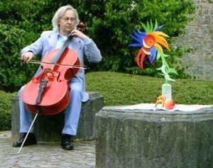 Ein Cellist spielt in einem Garten, neben ihm steht eine bunte Blume aus Papier.